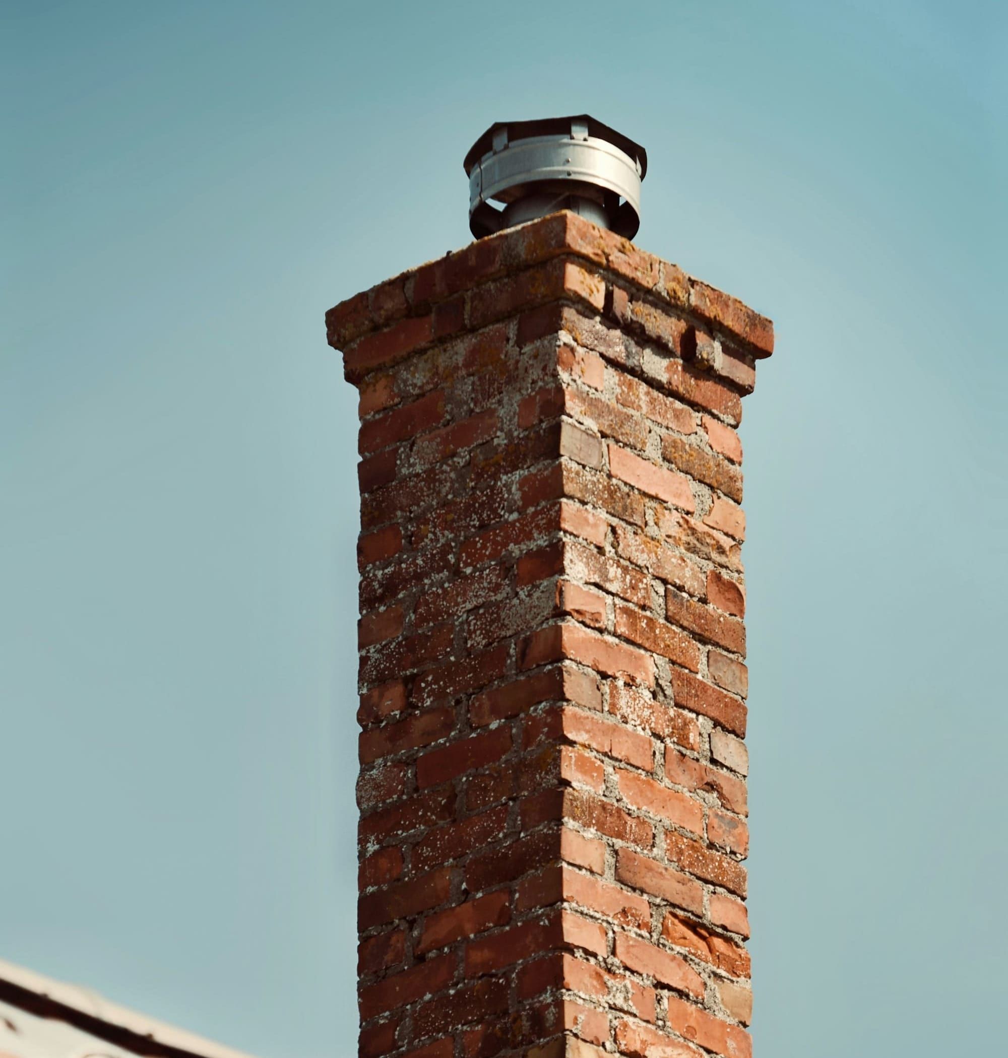 Weathered red brick chimney with a corbelled top and a round metal cap with a lid and open sides fitted over the flue
