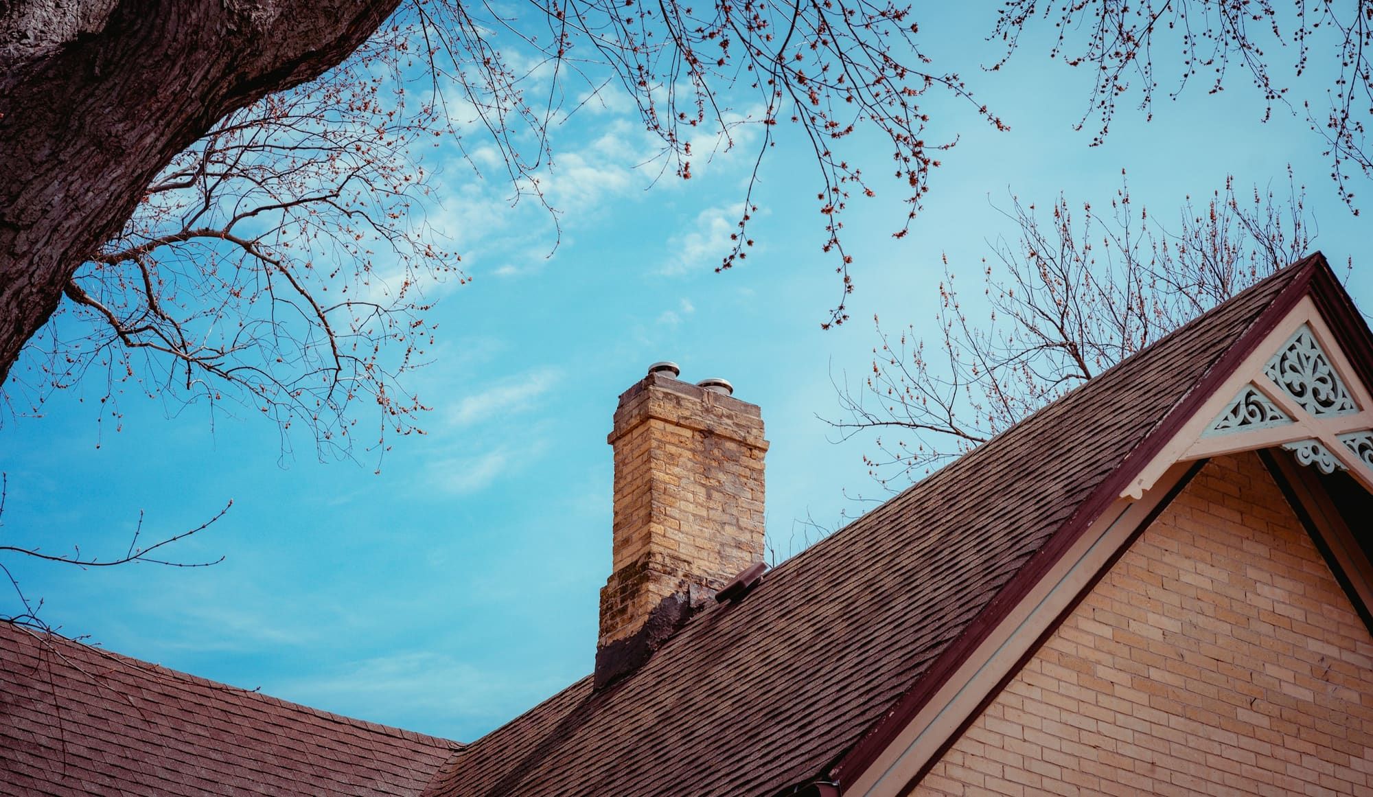 Buff brick chimney rising through an asphalt shingle roof beside a brick gable with decorative trim, under bare spring branches