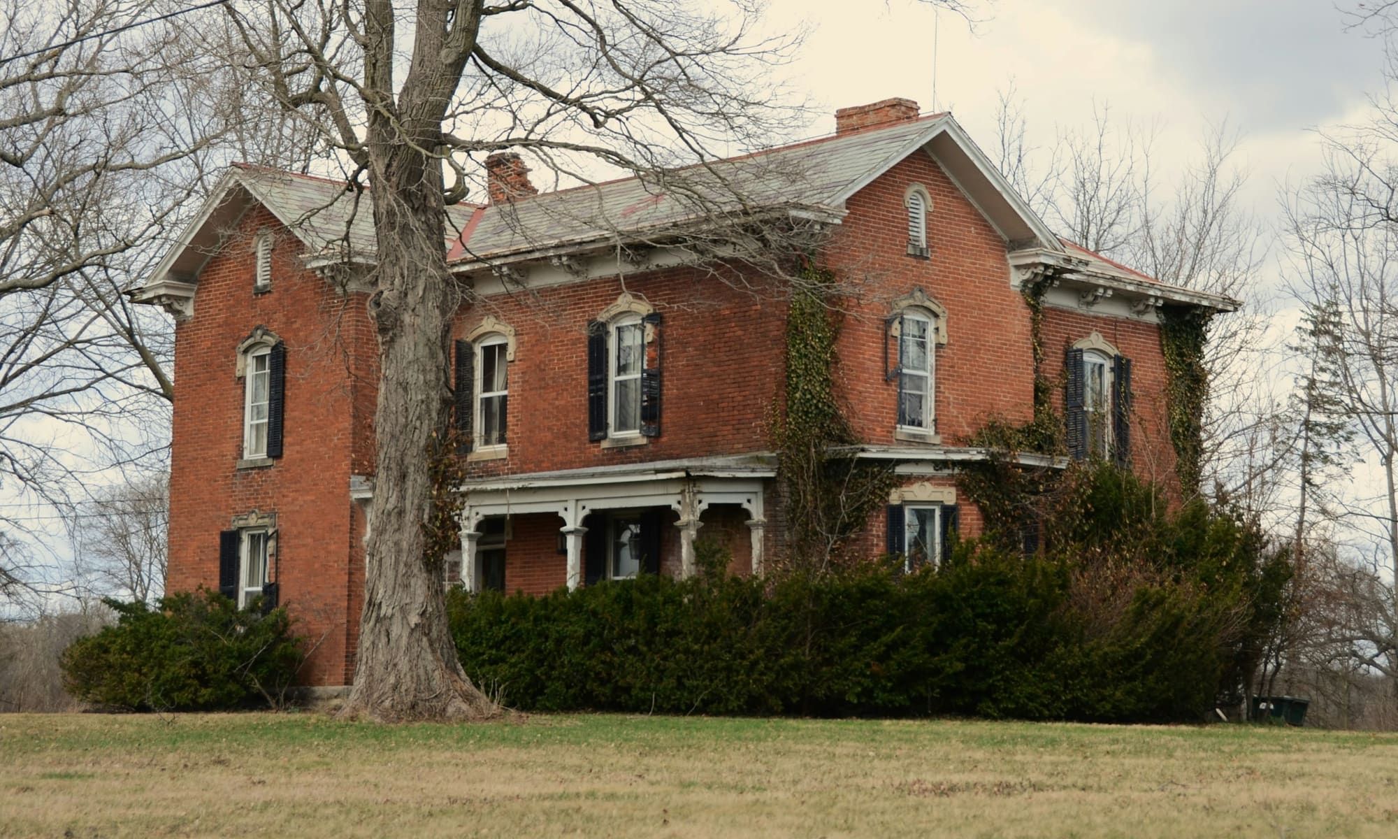 Old two-story red brick farmhouse with brick chimneys, a front porch and ivy on one corner, behind a large bare tree