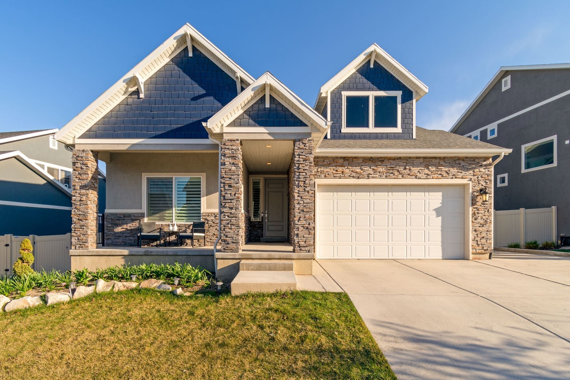 Front of a newer two-story house with stone veneer on the porch columns, entry and garage wall below dark blue shingle-sided gables