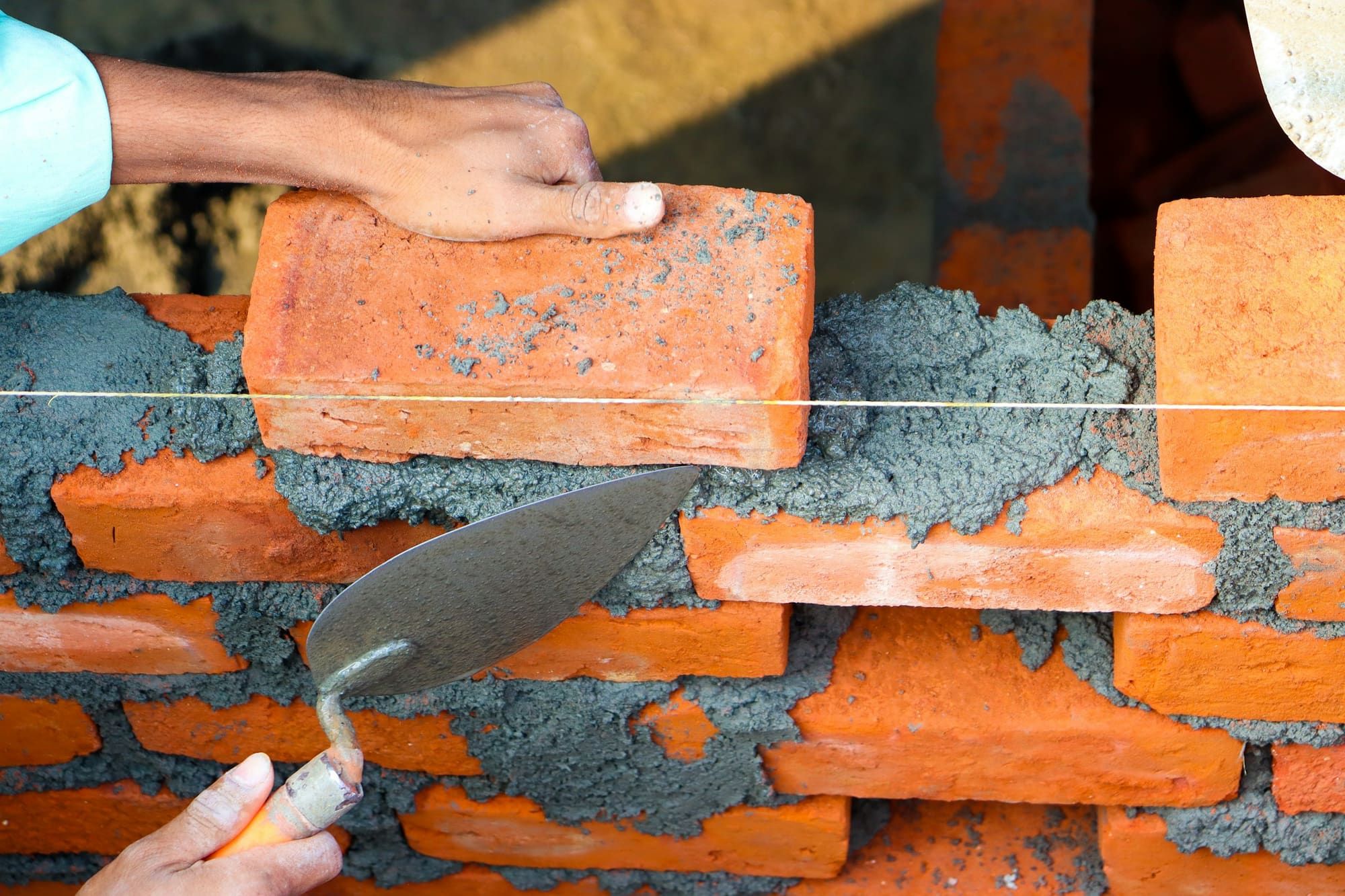 Hands setting a clay brick into a fresh bed of mortar along a string line, with a pointed brick trowel in the foreground