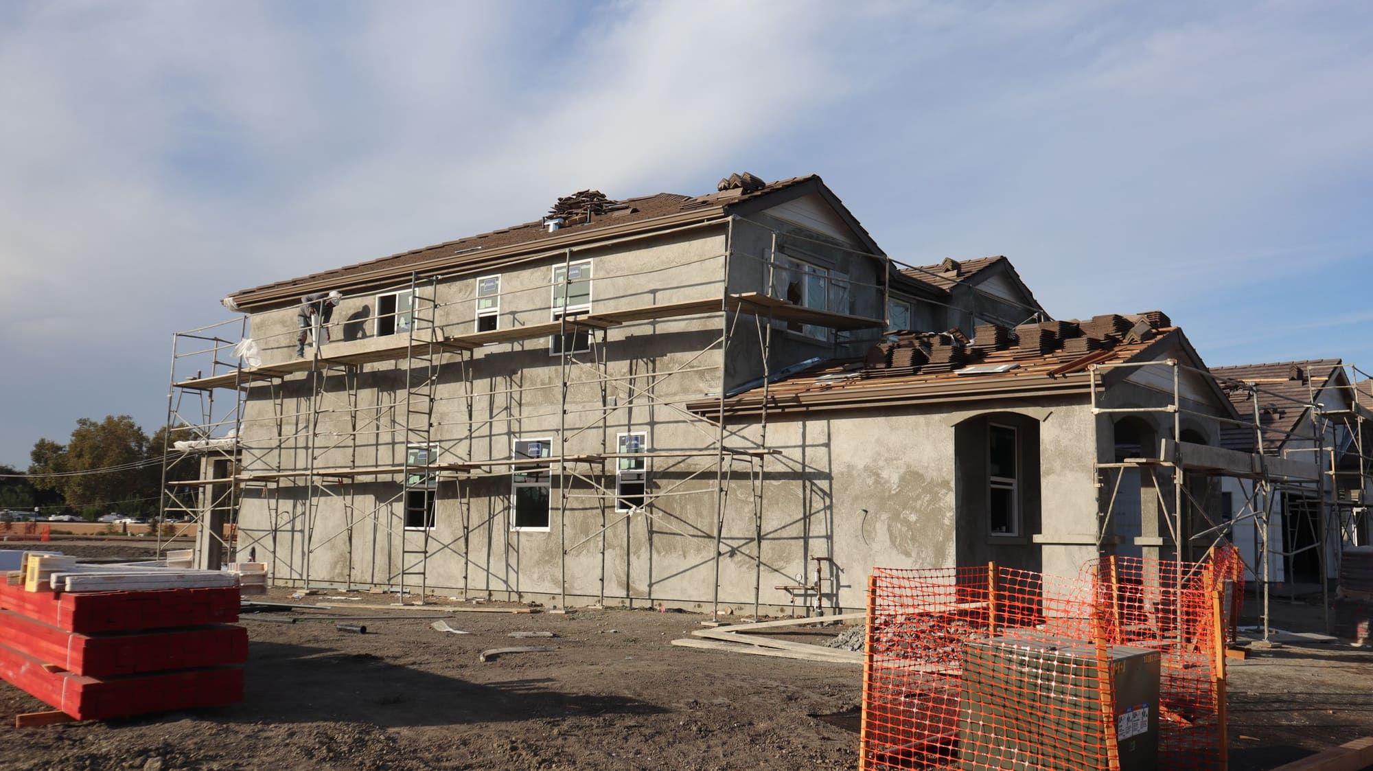 Two-story house under construction with a gray cement stucco base coat on every wall, frame scaffolding along the side and a worker on the top platform