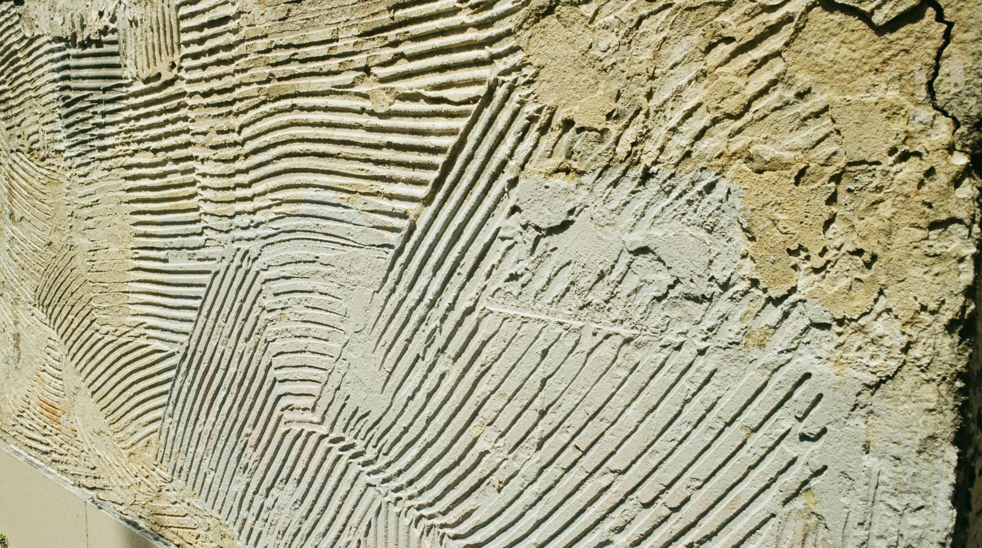 Close-up of a cement base coat on a wall raked into rows of horizontal and diagonal grooves, with a rougher tan coat beside it