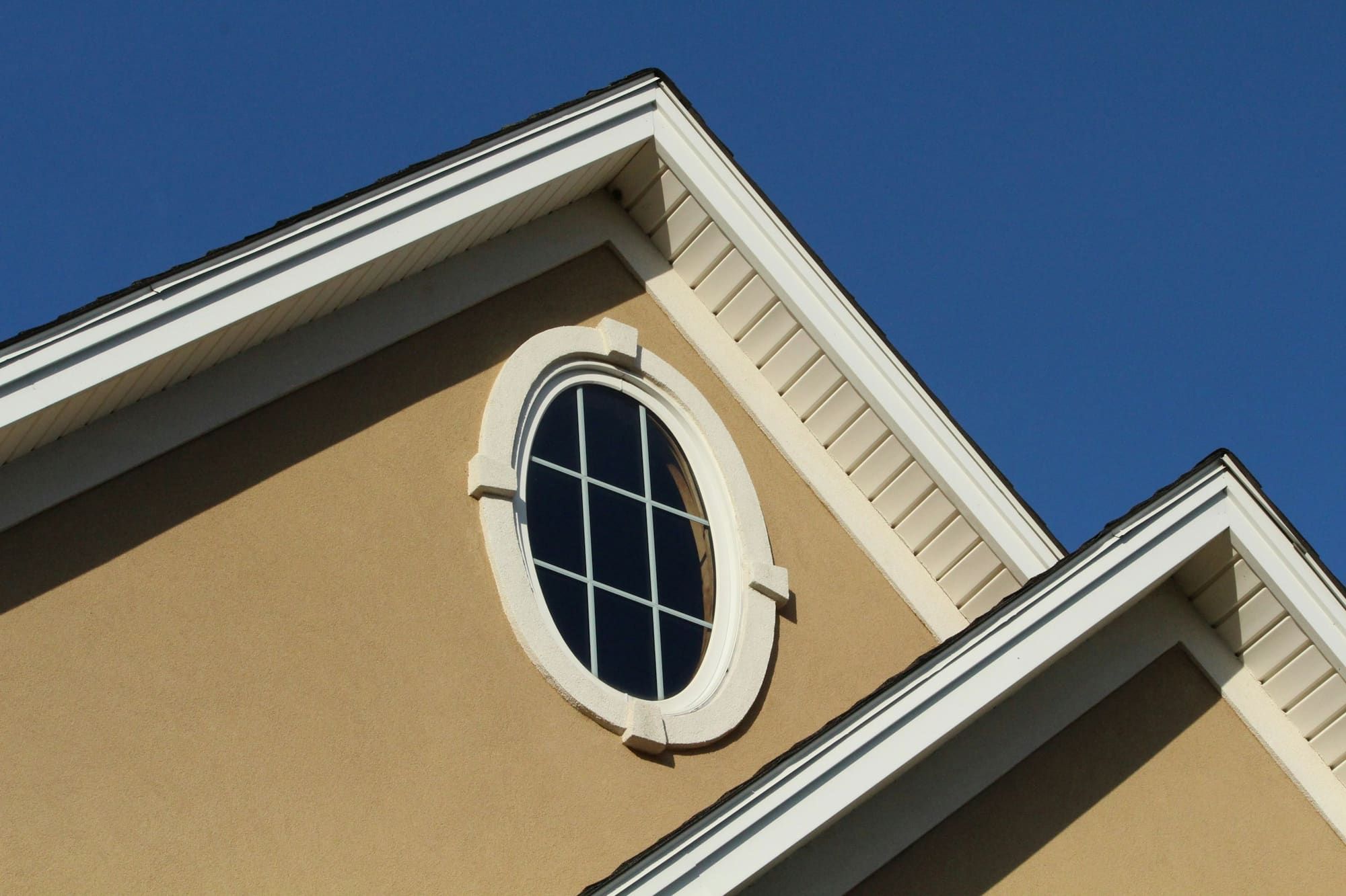 Tan smooth-finish stucco gable with an oval window framed by a raised cream trim band with keystone blocks, under white soffits and a deep blue sky