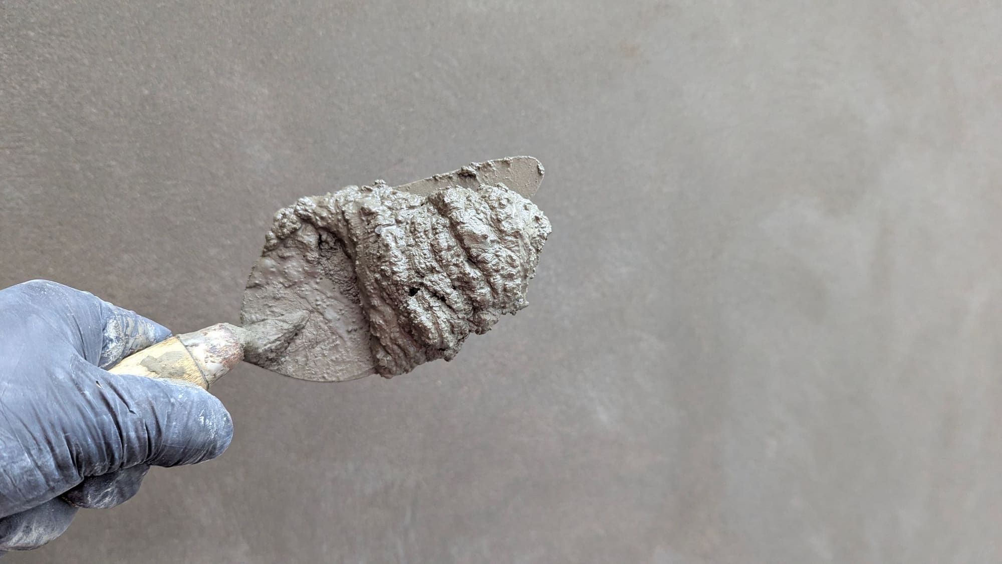 Gloved hand holding a pointed trowel loaded with wet gray cement plaster in front of a freshly floated gray wall